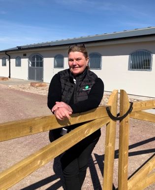 Kvinna står vid ett stall. Hon lutar sig mot ett staket. 